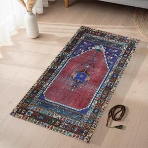 Può includere: Un tappeto rettangolare dal design tradizionale, con un campo centrale rosso incorniciato da un bordo blu. Il tappeto presenta motivi intricati nelle tonalità del rosso, blu e verde. Un set di grani di preghiera è appoggiato accanto al tappeto su un pavimento in legno chiaro.