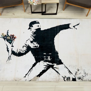 May include: A black and white rug featuring a stencil of a person throwing a bouquet of flowers. The person is wearing a black mask and a black cap. The background is a white wall with a distressed look.