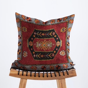 May include: A decorative pillow with a red, black, and gray patterned design. The pillow features a central geometric design with floral accents in orange, yellow, and green. The pillow is placed on a wooden stool.