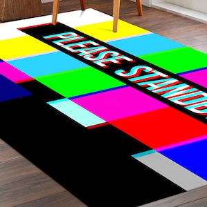 May include: A rectangular rug with a vibrant, multi-coloured design. The rug displays a pattern of horizontal bars in colours such as white, yellow, blue, green, red, and black. The words "PLEASE STANDBY" are printed diagonally across the rug.