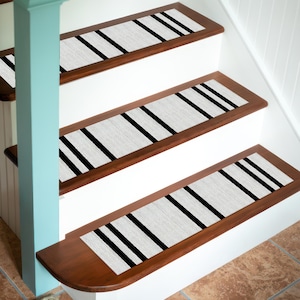 Può includere: Gradini con sfondo grigio chiaro e tre audaci strisce nere. I gradini sono posizionati su gradini in legno con un bordo marrone scuro e alzatine bianche. Il design si ripete su più gradini.