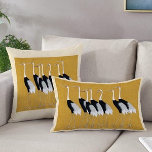 Puede incluir: Dos almohadas decorativas con fondo amarillo y un patrón de grúa blanco y negro. Las almohadas están en un sofá beige.
