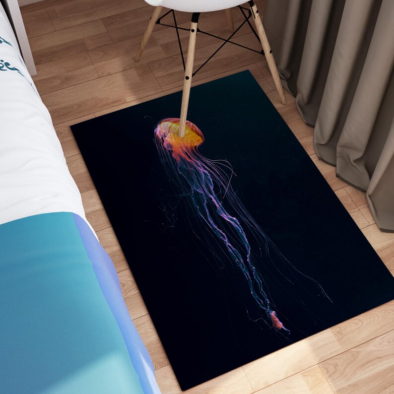 K&ouml;nnte beinhalten: Rechteckiger Teppich mit einem Quallen-Design auf dunklem blauem Hintergrund. Die Qualle hat eine gelb-orangefarbene Glocke und lange, flie&szlig;ende Tentakel in Lila und Rosa. Der Teppich liegt auf einem Holzboden, mit einem Stuhlbein sichtbar.