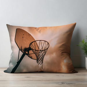 Puede incluir: Un cojín decorativo con una canasta de baloncesto contra un cielo nublado y anaranjado. El aro y el tablero están desgastados, con un acabado de metal oscuro. El cojín está sobre una superficie de madera.