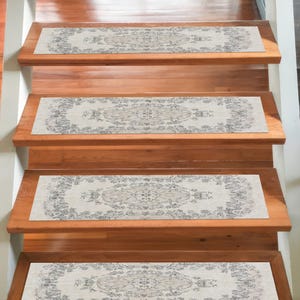 May include: Wooden staircase with four steps, each adorned with a rectangular rug. The rugs feature a cream-colored background with a detailed, symmetrical floral and geometric pattern in shades of gray and black.