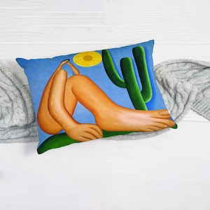 May include: A blue pillow with a painting of a person sitting with their legs crossed, a yellow sun, and a green cactus. The person is wearing a blue shirt and has brown hair.