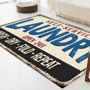 May include: A vintage-style laundry sign rug with a white background, black border, and blue text that reads "SELF-SERVICE LAUNDRY OPEN 24H WASH-DRY-FOLD-REPEAT".