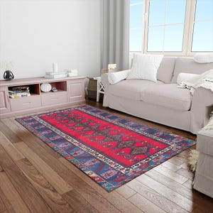 May include: A rectangular red rug with a central geometric pattern and blue borders. The rug is placed on a hardwood floor in a living room setting, with a sofa and window visible in the background.