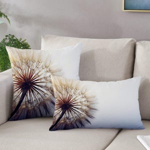 May include: Two throw pillows with a dandelion design. The pillows are white with a dandelion in the center. The dandelion is a light brown color with a dark brown center. The pillows are on a light brown couch.