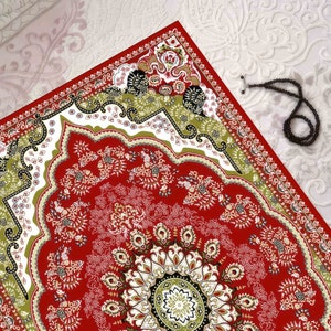 Puede incluir: Una alfombra de oración roja con un patrón geométrico verde, blanco y negro. La alfombra tiene un medallón central con un diseño floral.
