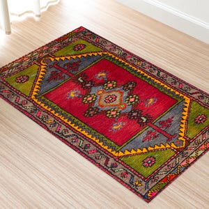 Puede incluir: Una alfombra rectangular con un campo central rojo, bordeada por patrones geométricos grises, verdes y amarillos. La alfombra presenta intrincados diseños florales y geométricos en varios colores, creando una estética vibrante y detallada.
