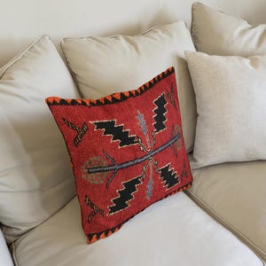Peut inclure: Un coussin rouge décoratif avec un motif géométrique noir, orange et bleu. Le coussin rectangulaire a un aspect texturé et repose sur un canapé crème avec d'autres coussins.