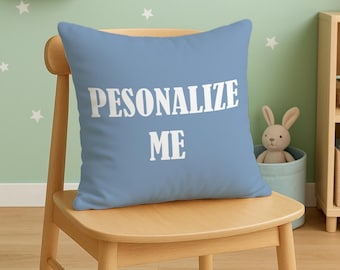 May include: A light blue square pillow with the words "PESONALIZE ME" in white block letters. The pillow is placed on a light wooden chair in a child's room. The room has a light green wall with white star decals.