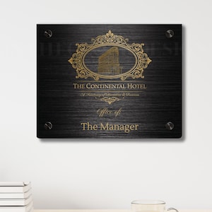 May include: A black and gold sign for The Continental Hotel, mounted on a wall. The sign features ornate gold detailing and the text "The Continental Hotel" and "Office of The Manager".
