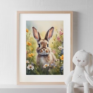 Può includere: Una stampa ad acquerello incorniciata di un coniglio marrone in un campo di fiori selvatici. Il coniglio ha lunghe orecchie ed è al centro dell'immagine. I fiori sono gialli, rosa e bianchi. Un coniglietto di peluche bianco è a destra.