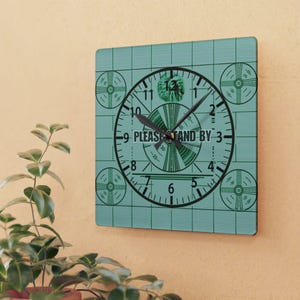 May include: A teal blue square clock with a black clock face and hands. The clock face has a green circle in the center with the text "PLEASE STAND BY" and a green and white design. The clock face also has black numbers 1 through 12 around the outside.