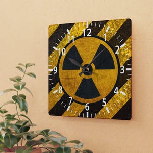 May include: Square wall clock with a yellow and black hazard stripe design. The clock face features a yellow radiation symbol with black hands and white numerals. The clock is mounted on a beige wall.