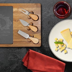 May include: A wooden cheese board with a slate insert featuring the text "The Continental Hotel". Includes three cheese knives, a plate of cheese, a glass of red wine, and a red napkin. The board is on a dark surface.