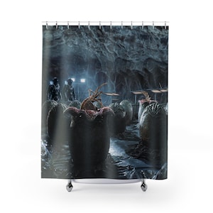 May include: Shower curtain featuring a science fiction scene. The image depicts a dark cave with several large, dark, egg-like structures. Two figures in space suits stand in the background. The scene is lit with a cool, blue light, creating a dramatic atmosphere.