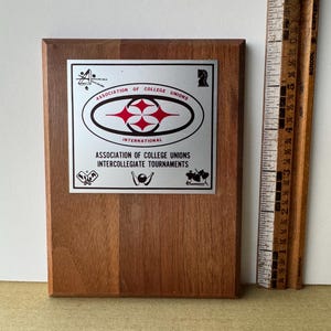 May include: A wooden plaque with a silver metal plate featuring the Association of College Unions International logo. The logo is a red and white stylized starburst design inside a black circle. The text "Association of College Unions International" and "Intercollegiate Tournaments" is printed on the plate.
