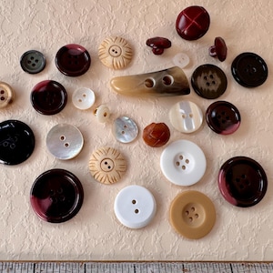 May include: A collection of assorted buttons in various shapes, sizes, and colors including black, white, brown, and burgundy. The buttons feature different textures, finishes, and hole counts, displayed alongside a ruler showing imperial measurements.
