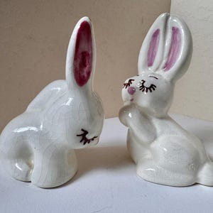 May include: Two white ceramic rabbit figurines with a glossy crackle glaze finish. Each bunny features painted pink inner ears, a small pink nose, and black eyelashes. One rabbit is positioned looking downward, while the other sits upright.