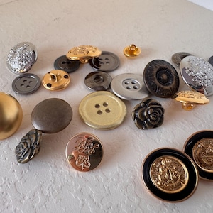 May include: A collection of assorted sewing buttons in various sizes, shapes, and finishes, including metallic gold, silver, bronze, and black tones. Some buttons feature intricate embossed patterns, floral designs, or crests, while others are plain.