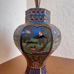May include: A cloisonné jar with a lid, featuring a landscape design with mountains, trees, and birds in blue, green, and white enamel on a brass base. A wooden ruler at the base shows the jar stands approximately 5.5 inches in total height.