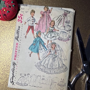 May include: A vintage Simplicity printed pattern for a doll, size 18 inches. The pattern features illustrations of various doll outfits, including dresses and a bridal gown. A red pin cushion and scissors are also visible.