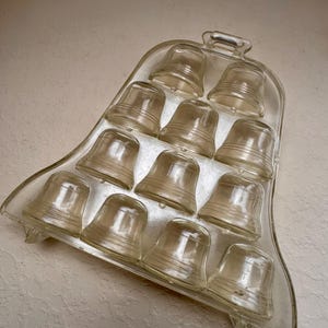 May include: A clear glass mold shaped like a large bell, featuring ten smaller bell-shaped indentations arranged in a triangular pattern. The mold has a smooth, transparent surface with a small handle at the top and two small feet at the base.