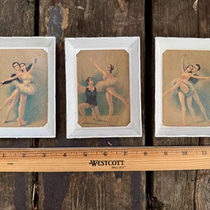 May include: Three framed vintage ballet prints. Each print features a couple in ballet poses, with the dancers in classic attire. The frames are white with a textured border. A wooden ruler is at the bottom, measuring up to 12 inches.