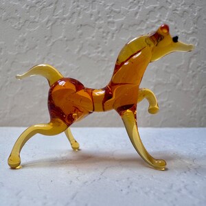 May include: A handcrafted glass horse figurine in shades of amber and yellow. The horse is posed mid-stride, with delicate legs and a flowing mane and tail. The figurine is a decorative item.