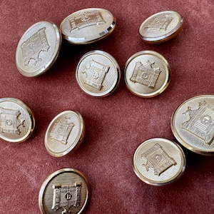 May include: A collection of gold-colored metal buttons with a detailed embossed design. The buttons are round and feature a central emblem surrounded by a textured border. The buttons are scattered on a burgundy surface.