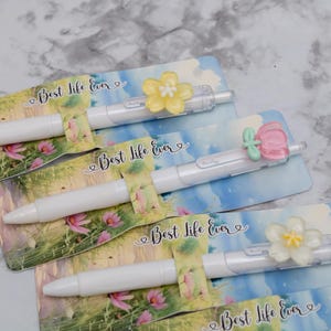 May include: White pens with floral embellishments and the text "Best Life Ever" on decorative cards. The pens have a clear barrel and are topped with yellow, pink, and white flower designs.