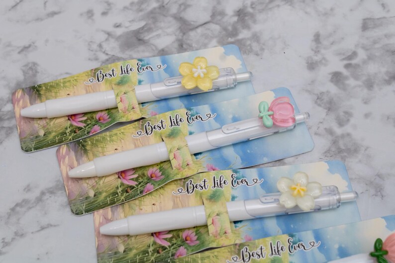 May include: White pens with floral embellishments and the text "Best Life Ever" on decorative cards. The pens have a clear barrel and are topped with yellow, pink, and white flower designs.