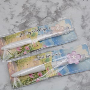 May include: Two white pens with pink flower accents, packaged on cards with landscape art and text. One card reads "No Hay Vida Mejor", the other "Best Life Ever".