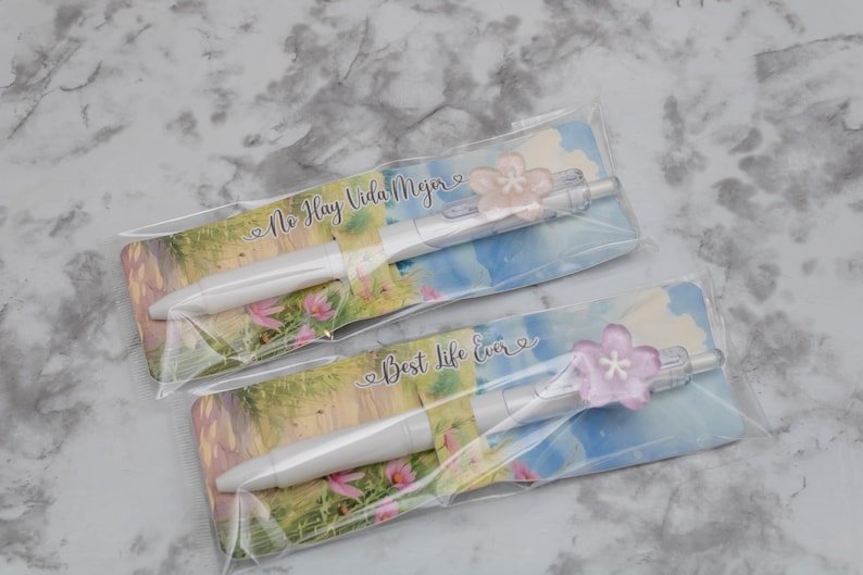 May include: Two white pens with pink flower accents, packaged on cards with landscape art and text. One card reads "No Hay Vida Mejor", the other "Best Life Ever".