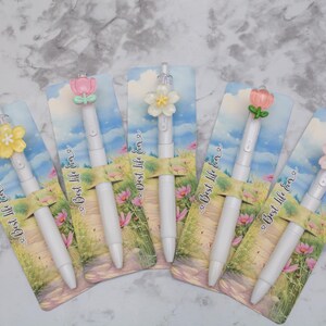 May include: Five white pens with flower-shaped toppers in various colors, including pink, yellow, and white. Each pen is displayed on a card with a landscape design and the text "Best life for".