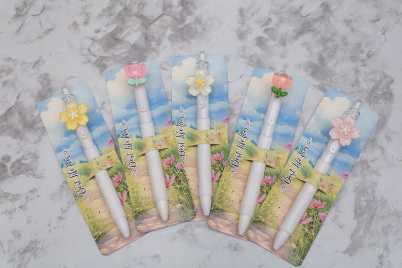 May include: Five white pens with flower-shaped toppers in various colors, including pink, yellow, and white. Each pen is displayed on a card with a landscape design and the text "Best life for".