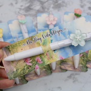 May include: White pens with flower-shaped toppers are displayed on cards with a landscape design and the text "No Hay Vida Mejor". The pens have different colored flower accents, including yellow, pink, and green.