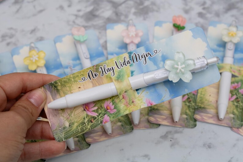 May include: White pens with flower-shaped toppers are displayed on cards with a landscape design and the text "No Hay Vida Mejor". The pens have different colored flower accents, including yellow, pink, and green.