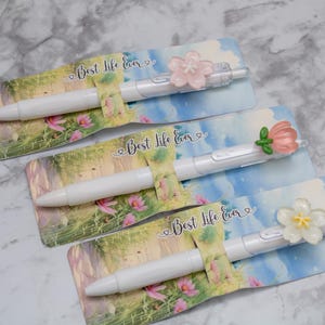 May include: Three white pens with floral accents and the text "Best Life Ever" on decorative cards. Each pen features a different flower design in pink, white, and green. The cards have a watercolor-style landscape background.