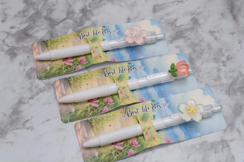 May include: Three white pens with floral accents and the text "Best Life Ever" on decorative cards. Each pen features a different flower design in pink, white, and green. The cards have a watercolor-style landscape background.