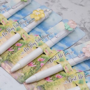 May include: White pens with floral accents and the text "No Hay Vida Mejor" and "Best Life Ever" on the packaging. The pens have decorative flower toppers in various colors, set against a floral and sky-blue background.