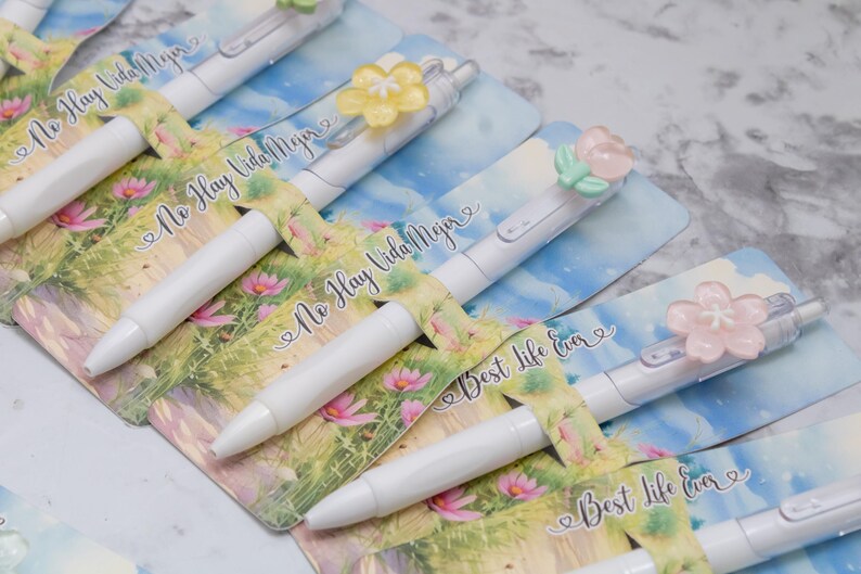 May include: White pens with floral accents and the text "No Hay Vida Mejor" and "Best Life Ever" on the packaging. The pens have decorative flower toppers in various colors, set against a floral and sky-blue background.