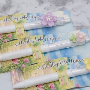 May include: White pens with flower accents rest on cards with a floral and sky design. The pens have a clear plastic clip and are topped with a purple, green, and yellow flower. The text "No Hay Vida Mejor" is printed on the cards.