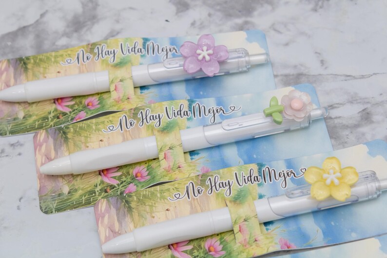 May include: White pens with flower accents rest on cards with a floral and sky design. The pens have a clear plastic clip and are topped with a purple, green, and yellow flower. The text "No Hay Vida Mejor" is printed on the cards.