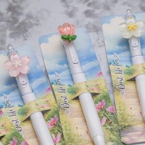 May include: White pens with flower-shaped toppers in pink, white, and peach colors. The pens are displayed on cards with a landscape design and the text "Best life ever". The pens are a stationery item.