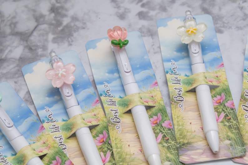 May include: White pens with flower-shaped toppers in pink, white, and peach colors. The pens are displayed on cards with a landscape design and the text "Best life ever". The pens are a stationery item.