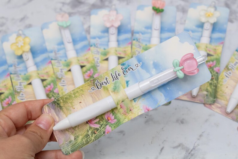 May include: White pens with flower-shaped toppers and decorative cards. The cards feature a watercolor design with the text "Best Life Ever". The pens have a smooth, white barrel and are ideal for gifts or stationery.